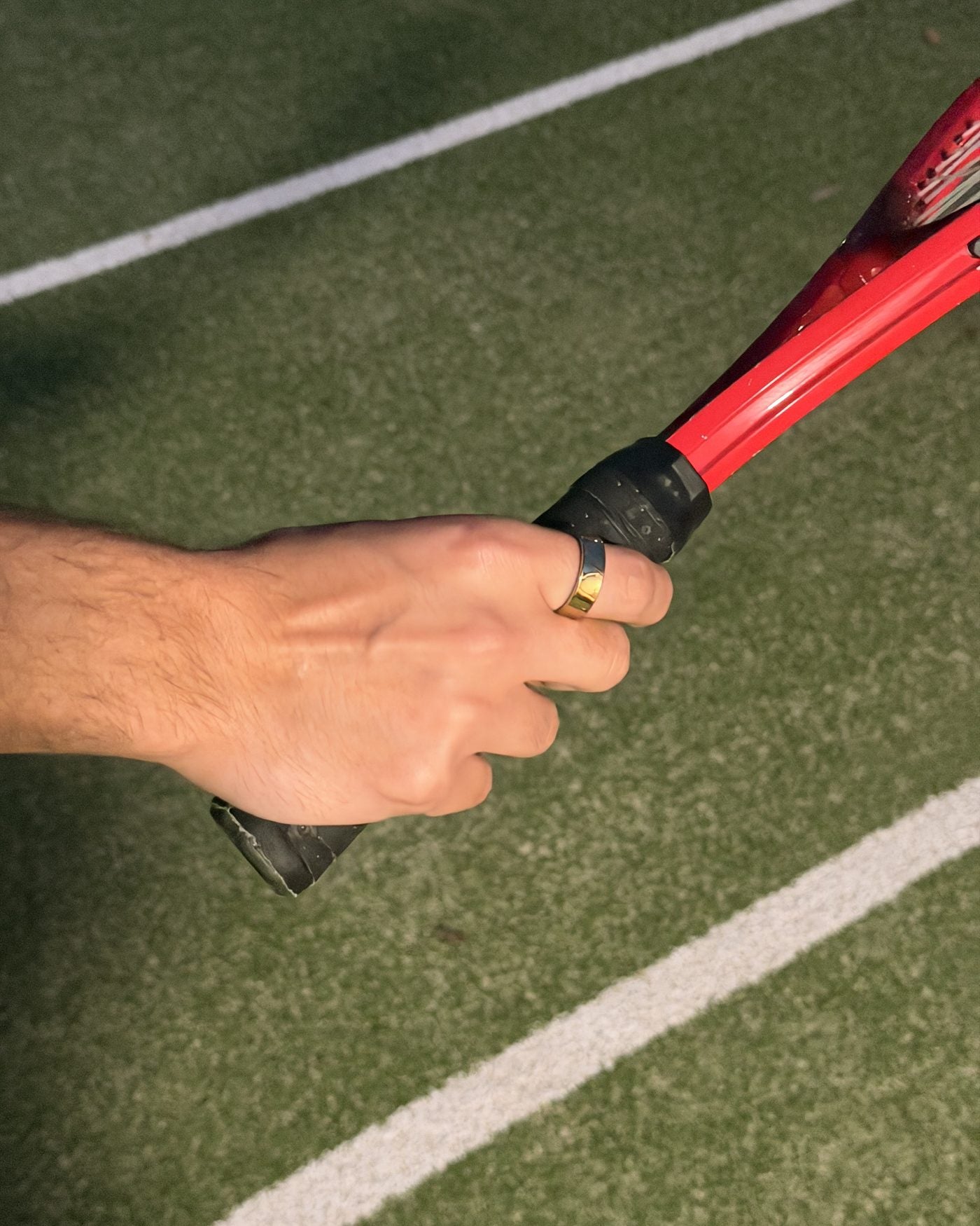 A hand wearing the gold Mudrika Ring gripping the handle of a red racquet on a green court