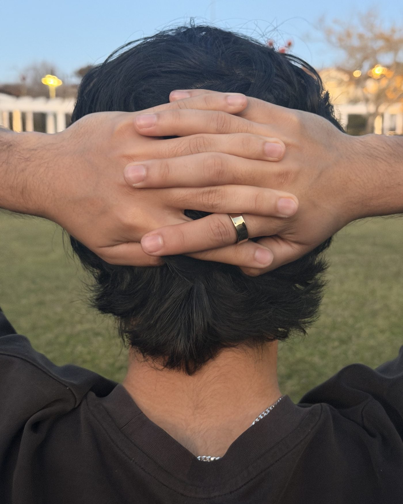 A person resting with both hands clasped behind their head at golden hour, the gold Mudrika Ring visible on one finger